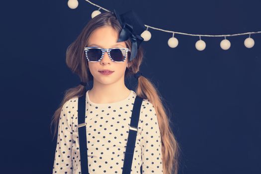 Fashion photo of young hipster child wearing cool clothes and sunglasses standing over dark wall, instagram toned image