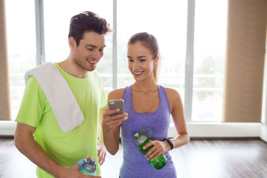fitness, sport, technology and slimming concept - smiling young woman and personal trainer with smartphone and water bottles in gym