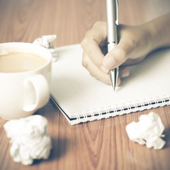 woman hand writing with pen on notebook.there are crumpled paper and coffee cup on wood table background vintage style