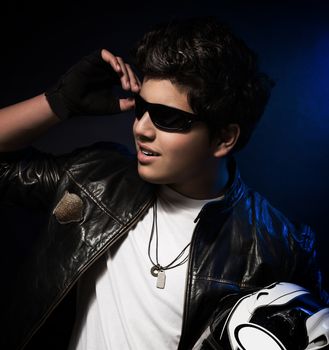 Portrait of handsome stylish teen boy posing in the studio over dark blue background, wearing bikers outfit and holding helmet in hands
