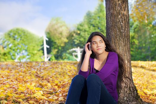 Teen girl sitting against autumn tree using cell phone