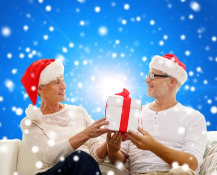 family, holidays, christmas, age and people concept - happy senior couple in santa helper hats with gift box over blue snowy background