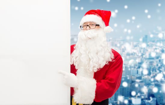christmas, holidays, advertisement, gesture and people concept - man in costume of santa claus pointing finger to white blank billboard over snowy city background