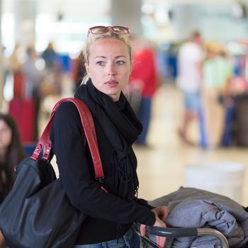 Casual blond young woman using her cell phone while queuing for flight check-in and baggage drop. Wireless network hotspot enabling people to access internet conection. Public transport.