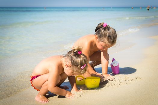 Two little girls playing with sand near the crystal clear sea.