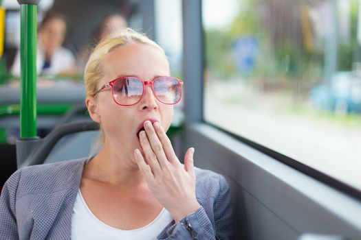 Blonde caucasian business woman yawning while traveling by bus early morning. Public transport and commuting to work.