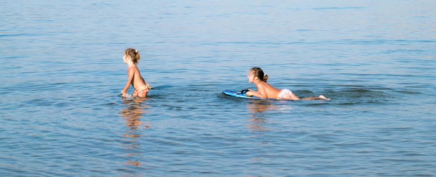 Two little girls playing in the sea with a blue floating board.