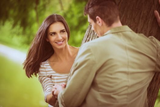 Beautiful girl in the park with her boyfriend enjoys playing hide behind a tree.