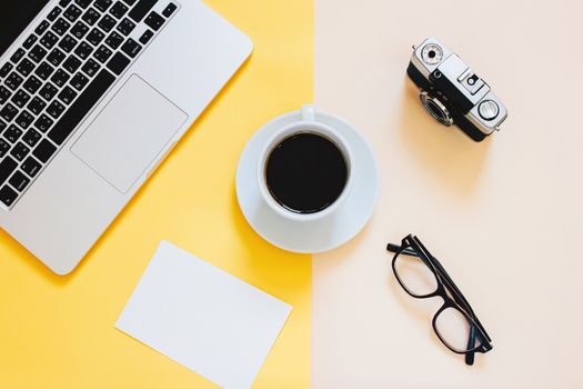 Creative flat lay photo of workspace desk with laptop, blank paper, coffee, eyeglasses and film camera on yellow modern background
