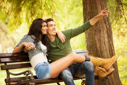 Attractive young heterosexual couple sitting on a park bench. Attractive girl sitting on the bench, and her long pretty legs holding on knees her boyfriend, who outstretched finger pointing at something.