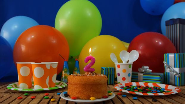 Birthday cake on rustic wooden table with background of colorful balloons, gifts, plastic cups and plastic plate with candies and blue wall in the background