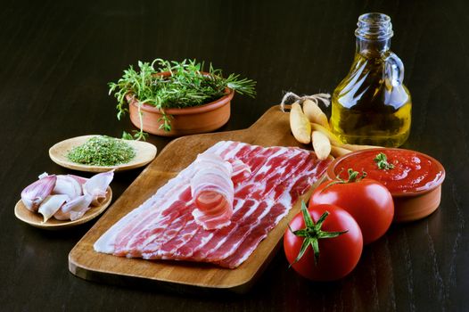 Pancetta on Wooden Cutting Board and Ingredients with Tomatoes, Garlic, Rosemary, Tomato Sauce, Bread Sticks, Spices and Olive Oil closeup on Dark Wooden background