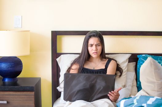 Biracial teen girl using laptop sitting in bed, disgusted expres