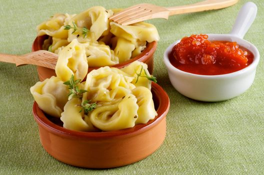 Arrangement of Delicious Meat Cappelletti with Herbs in Ceramic Bowls, Tomatoes Sauce and Wooden Forks closeup on Green Napkin background