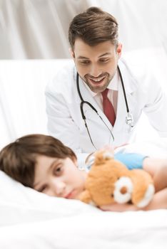 upset child patient lying in bed with smiling doctor behind