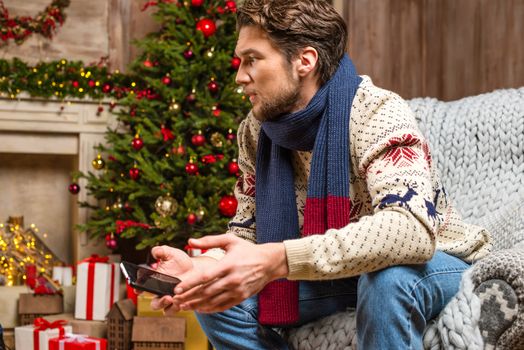 Handsome man in knitted sweater and scarf sitting in chair and holding smartphone in cozy room decorated for Christmas