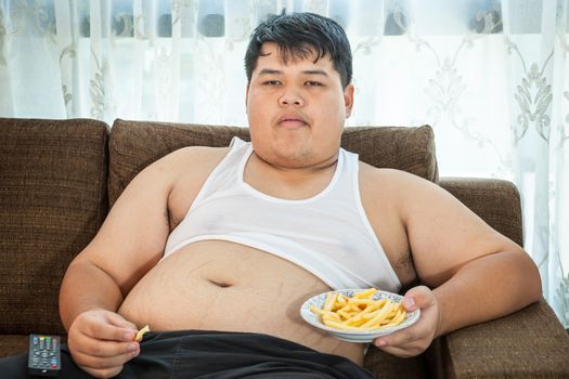 Lazy overweight asian male sitting with fast food on couch and watching television