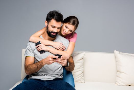 Young couple hugging on white couch and using smartphones 
