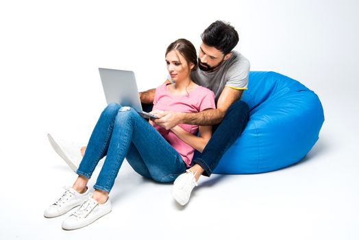 Young couple sitting on big blue pouf and looking at laptop, on white