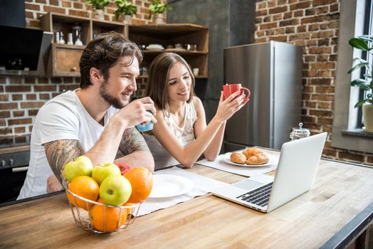 Young couple using laptop while having breakfast at kitchen table 