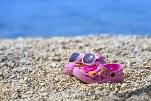 Pink beach kid's crocs / sandals and sunglasses on sandy beach.