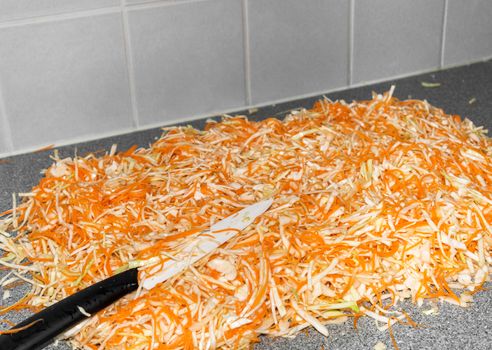 Homemade sauerkraut preparation - fermented cabbage and carrots, rustic winter food. Shredded cabbage, carrots, and knife on kitchen countertop.