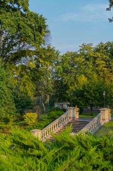 A staircase in a park with low handrails with lanterns for lighting in the evening is among trees and green lawns.
