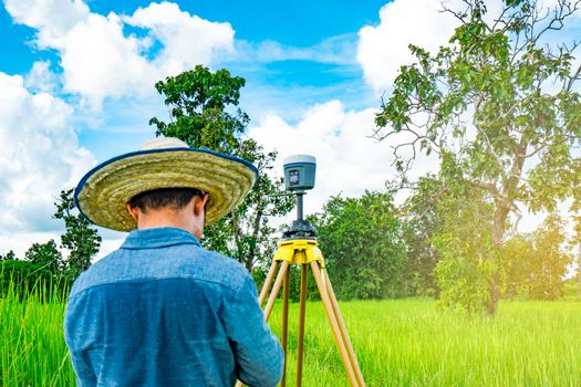 Asian smart engineer or surveyor in black jeans and long sleeve shirt and woven bamboo hat. He is working on controller screen for surveying land in rice field, Thailand. GPS surveying instrument.