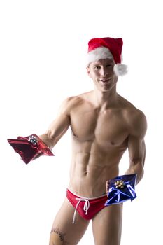 Athletic shirtless young man in Santa Claus hat standing holding Christmas gifts to celebrate the season, isolated on white looking at camera