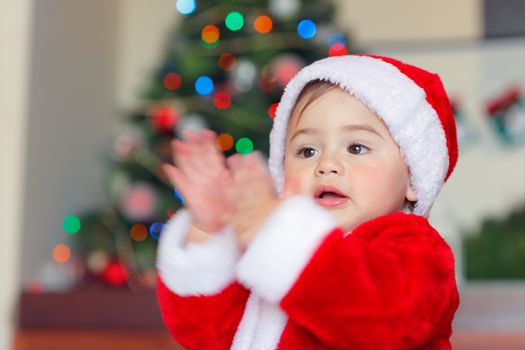 Portrait of a cute happy little boy having fun at home in Christmas eve, adorable child wearing Santa costume clapping near Christmas tree