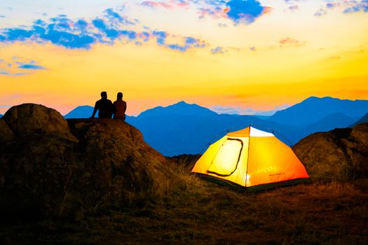 Young Couple Sitting on the Rock near Illuminated Tent and Watching the Beautiful Evening Mountain View with Sunset Sky