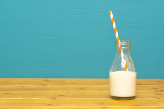 One-third pint glass milk bottle half full with fresh creamy milk with a retro paper straw, on a wooden table against a teal background