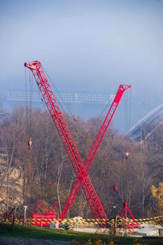 Construction process with construction cranes with red gibbets looks as letter X. Autumn fog and river bridge with big floating hotel on blurred background. Forest around cranes.
