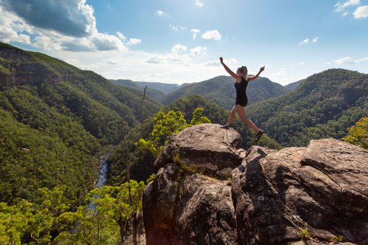 Girl leaping on high rocky cliff with mountain river backdrop