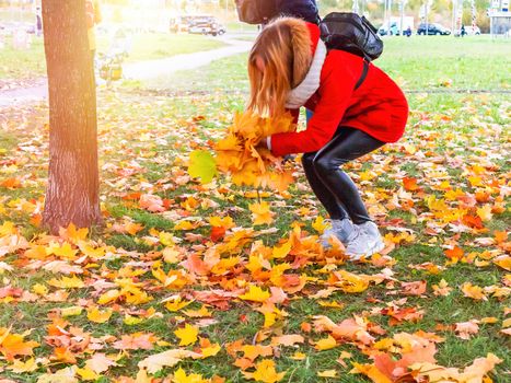 Young beautiful blonde girl in a red jacket enjoying a warm autumn day in the park and having fun, playing with autumn maple leaves