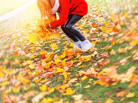 Young beautiful blonde girl in a red jacket enjoying a warm autumn day in the park and having fun, playing with autumn maple leaves