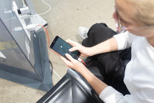 A young woman sitting at a charging station and looking at her smartphone. Recharging mobile phones from free charge station at the airport.