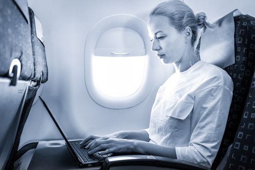 Attractive caucasian female passenger working at modern laptop computer using wireless connection on board of commercial airplane flight