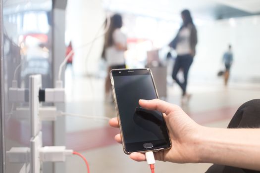 Female hands holding and using smartphone while charging it in a public place using electric plug and a charging cable