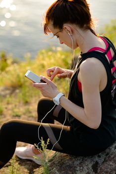 Young Woman Runner Using Smartphone and Listening to Music at Sunset on the Mountain Trail. Sports Concept.