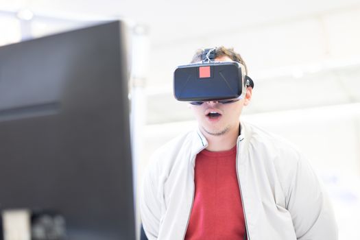 Young man wearing virtual reality headset and gesturing while sitting at his desk in creative office