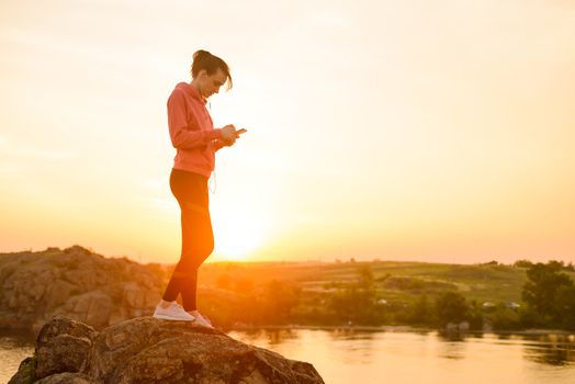 Woman Runner Resting after Workout, Using Smartphone and Listening to Music at Sunset on the Rock. Sports Concept.