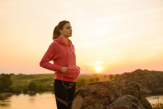 Woman Runner Resting after Workout and Listening to Music at Sunset on the Rock. Sports Concept.