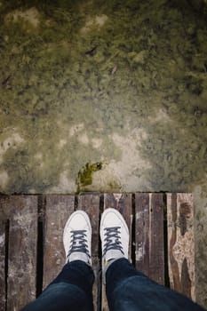 top view of female legs in white sneakers and jeans on the background of an old dirty autumn lake-river and a wooden platform. Copy space