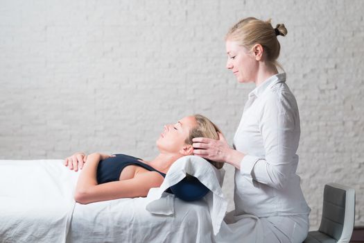 Professional female masseur giving relaxing massage treatment to young female client. Hands of masseuse on forehead of young lady during procedure of spa facial massage