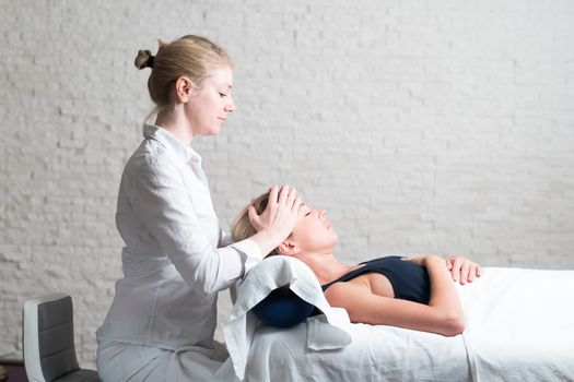 Professional female masseur giving relaxing massage treatment to young female client. Hands of masseuse on forehead of young lady during procedure of spa facial massage