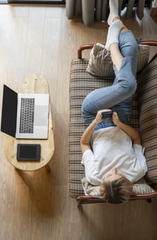 Woman use a smartphone while lying on a sofa with a laptop and ereader on a table. Studying online, freelance. Self employed or freelance girl use a phone and resting from work whith a notebook.