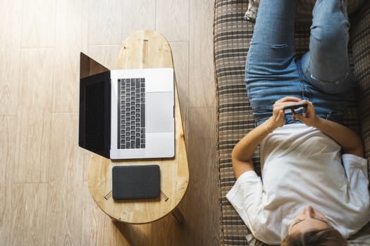 Woman use a smartphone while lying on a sofa with a laptop and ereader on a table. Studying online, freelance. Self employed or freelance girl use a phone and resting from work whith a notebook.