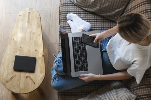 Woman with a laptop is sitting on a sofa and use a smartphone. Study and work online, freelance. Self employed girl is working with her notebook sitting on a couch with a phone and ereader on table.