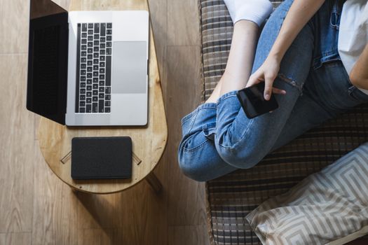 Woman with a laptop is sitting on a sofa and use a smartphone. Study and work online, freelance. Self employed girl is working with her notebook sitting on a couch with a phone and ereader on table.